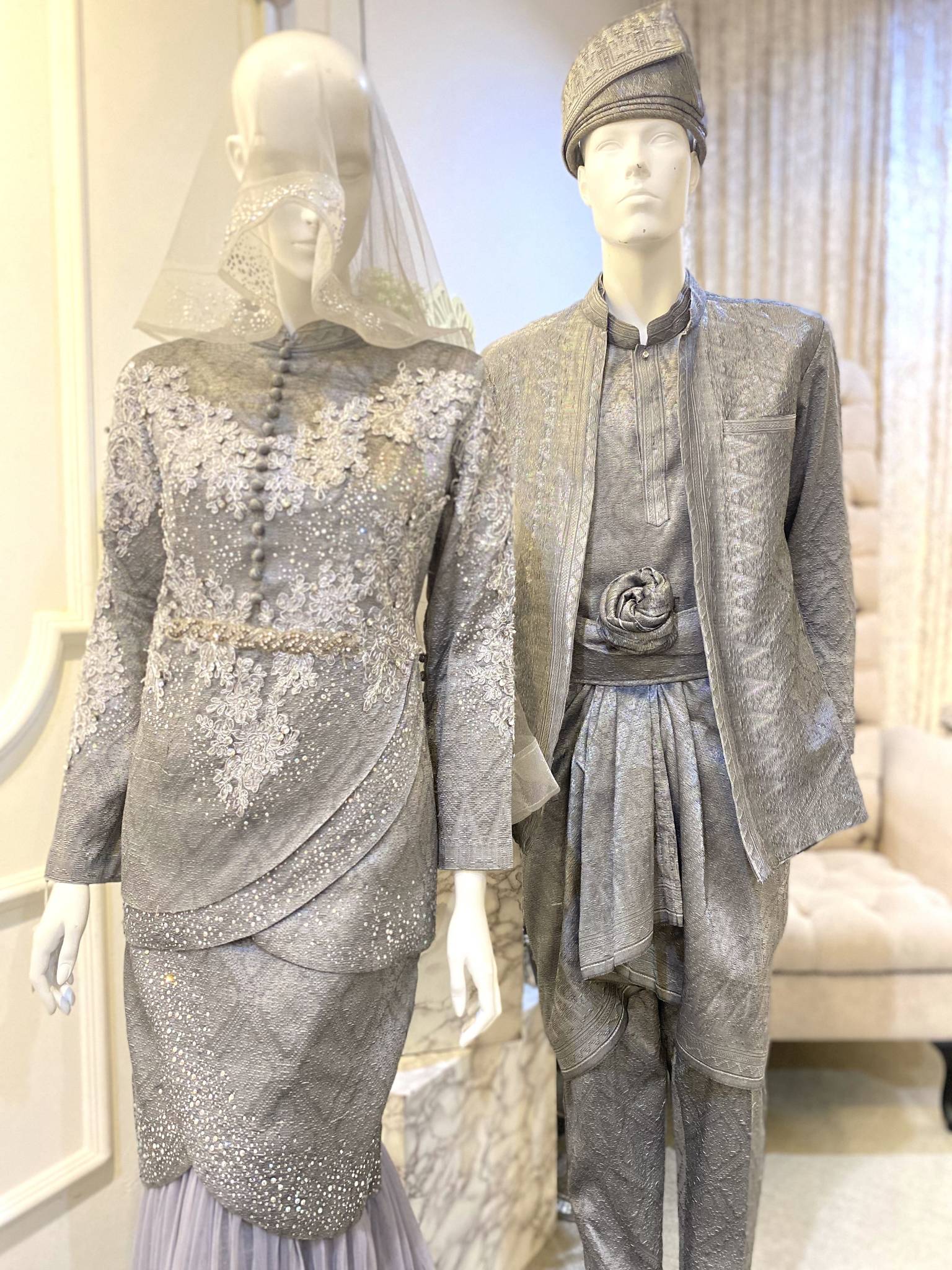 Two-piece Malay wedding dress in songket fabric in dark grey and silver. The top is a tulip-style top with a sweetheart neckline and off-the-shoulder sleeves. The skirt is a full-length skirt with a slit up the side. The dress is embellished with delicate silver embroidery.