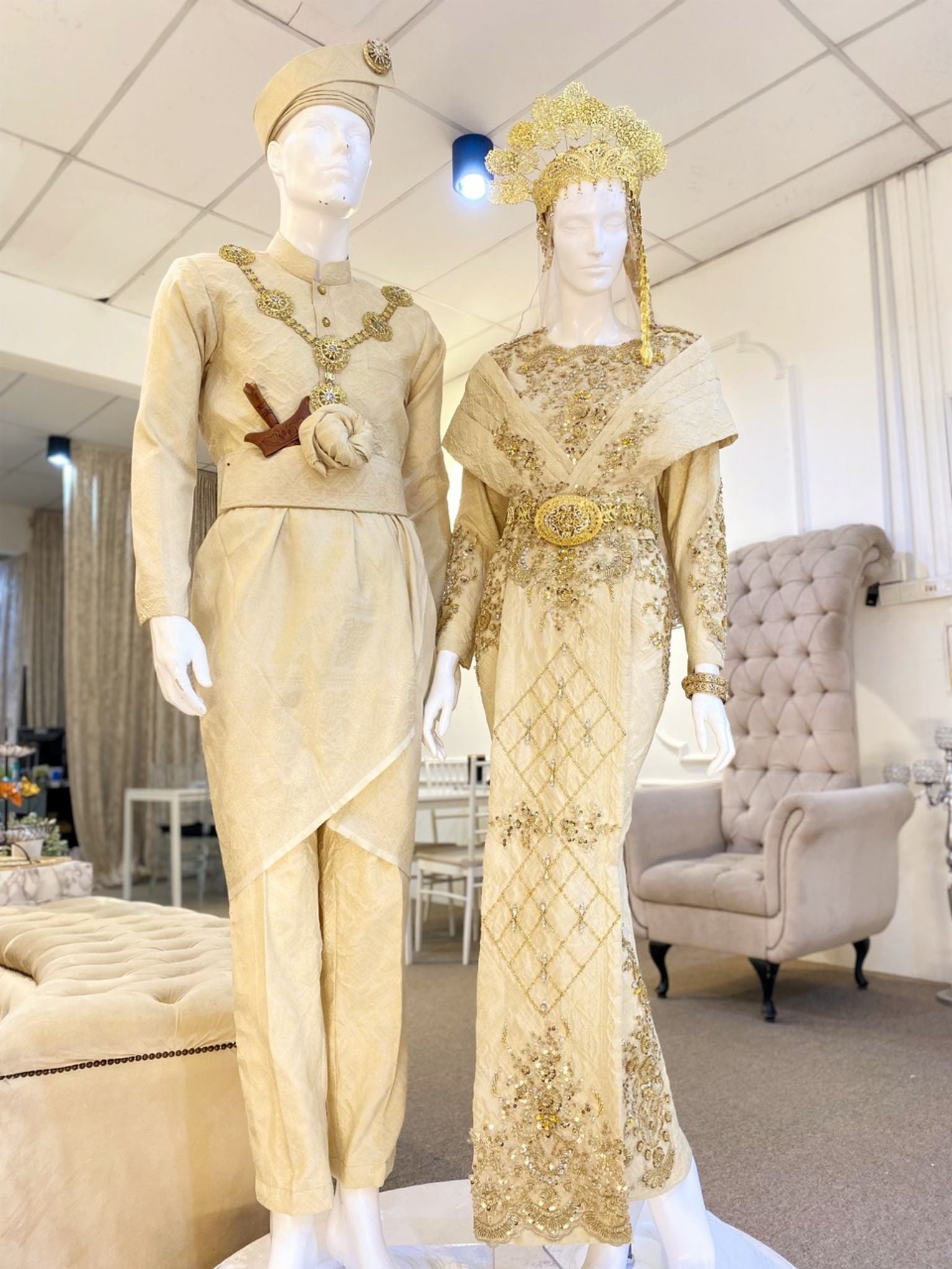 This image shows a man and a woman mannequin wearing traditional Malay wedding attire. The man is wearing a kot raihan, while the woman is wearing a dress. The mannequins are standing next to each other, and they are both smiling. The image is perfect for a wedding website or a clothing store that rent baju sanding.