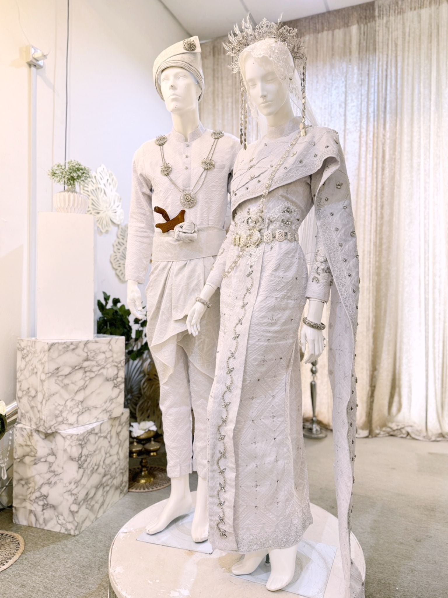 A photograph showcasing Nilam Sari's Baju Pengantin Songket in White with Silver, a blend of Thai-inspired bridal attire. The image captures the bride's ensemble, featuring a graceful Thai Dress design crafted from luxurious Songket 2D material, and the groom's attire, adorned with the intricate Kot Raihan 2pcs