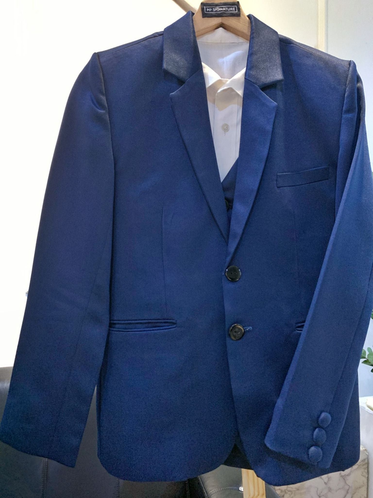 Blue suit jacket hanging on a wooden hanger. The jacket is made of a light blue fabric and has a single-breasted design with a notch lapel. It is suitable for formal occasions, such as weddings or job interviews.