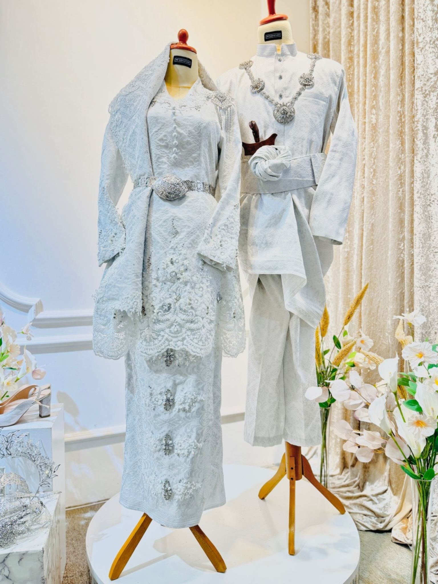 REMBULAN Baju Pengantin Kurung Songket in White with Silver, showcasing exquisite bridal and groom attire. The bride's ensemble features a stunning Kurung with Selempang, while the groom's attire displays the distinguished Kot Raihan 2pcs design. Crafted from luxurious Songket 2D material, available in sizes ranging from XS to M for the bride and S to L for the groom. Embrace tradition and elegance with this timeless bridal ensemble.