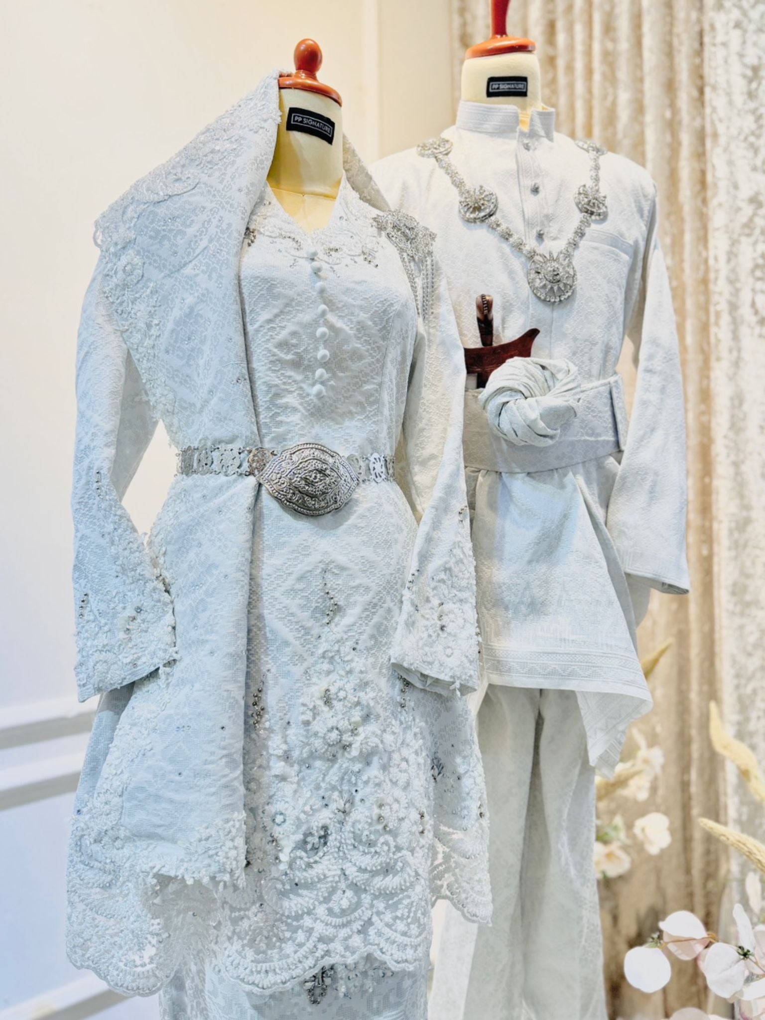 REMBULAN Baju Pengantin Kurung Songket in White with Silver, showcasing exquisite bridal and groom attire. The bride's ensemble features a stunning Kurung with Selempang, while the groom's attire displays the distinguished Kot Raihan 2pcs design. Crafted from luxurious Songket 2D material, available in sizes ranging from XS to M for the bride and S to L for the groom. Embrace tradition and elegance with this timeless bridal ensemble.